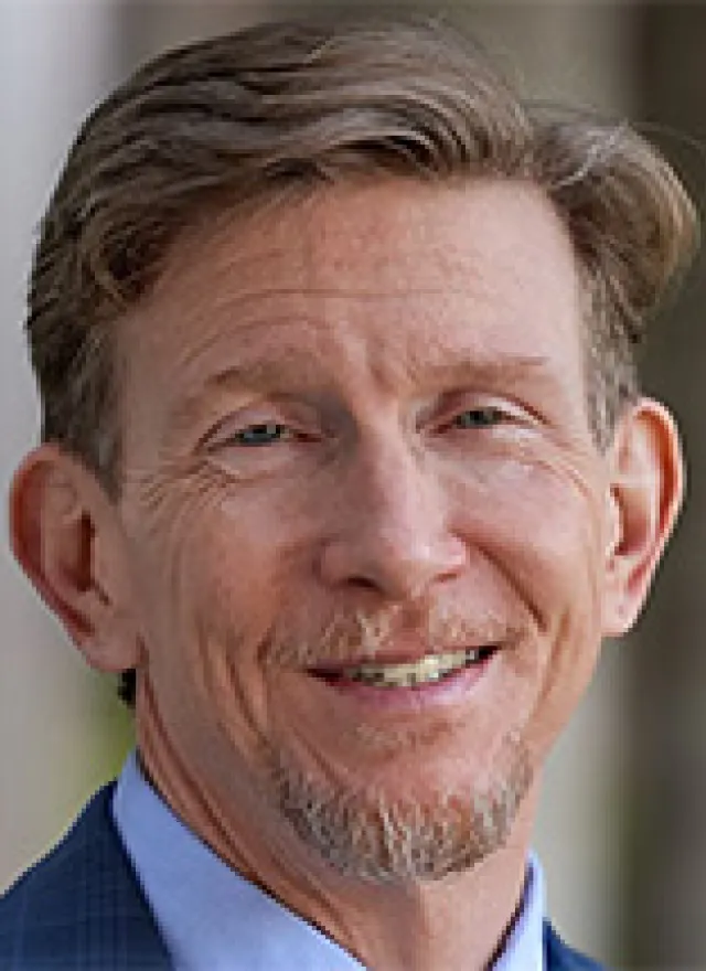 A middle-aged man with light brown hair, a short beard, and mustache is smiling and wearing a blue suit jacket and light blue shirt.