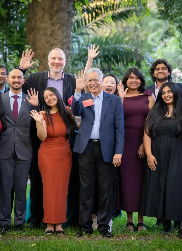 A group of people dressed in formal attire stand outdoors on grass, smiling and waving at the camera with trees in the background.