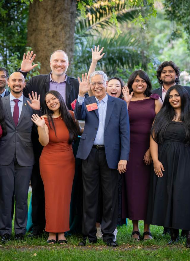 A group of people dressed in formal attire stand outdoors on grass, smiling and waving at the camera with trees in the background.