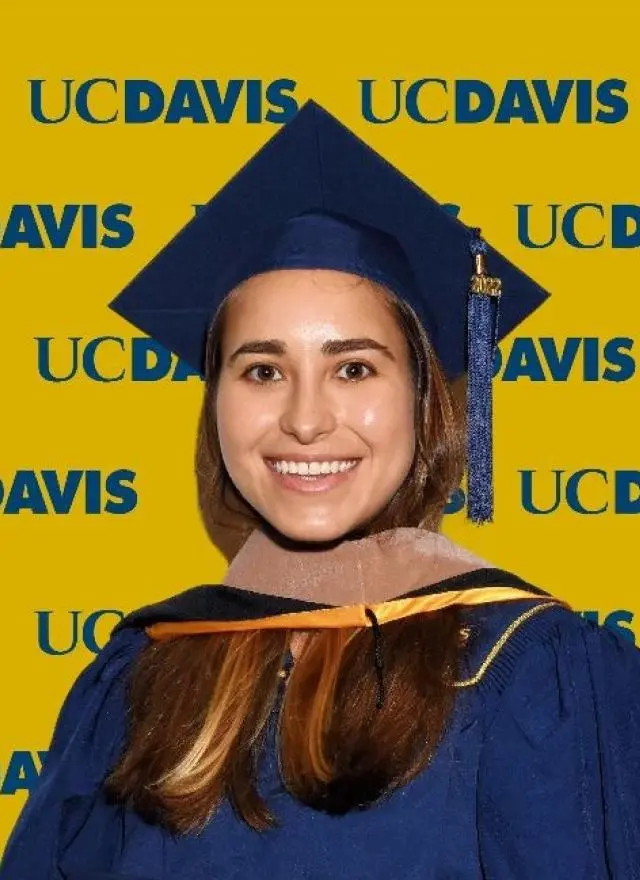Maria Ahumada wearing a blue graduation cap and gown stands in front of a yellow UC Davis backdrop with repeated logos.