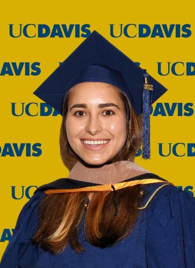Maria Ahumada wearing a blue graduation cap and gown stands in front of a yellow UC Davis backdrop with repeated logos.