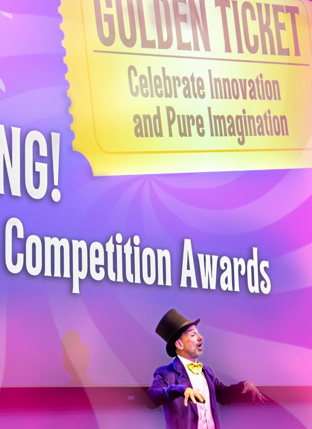A person dressed as Willy Wonka stands in front of a screen displaying “BIG BANG! Business Competition Awards” and “Golden Ticket: Celebrate Innovation and Pure Imagination.”