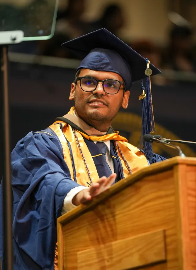 A graduate in cap and gown stands at a podium, addressing an audience during a commencement ceremony.
