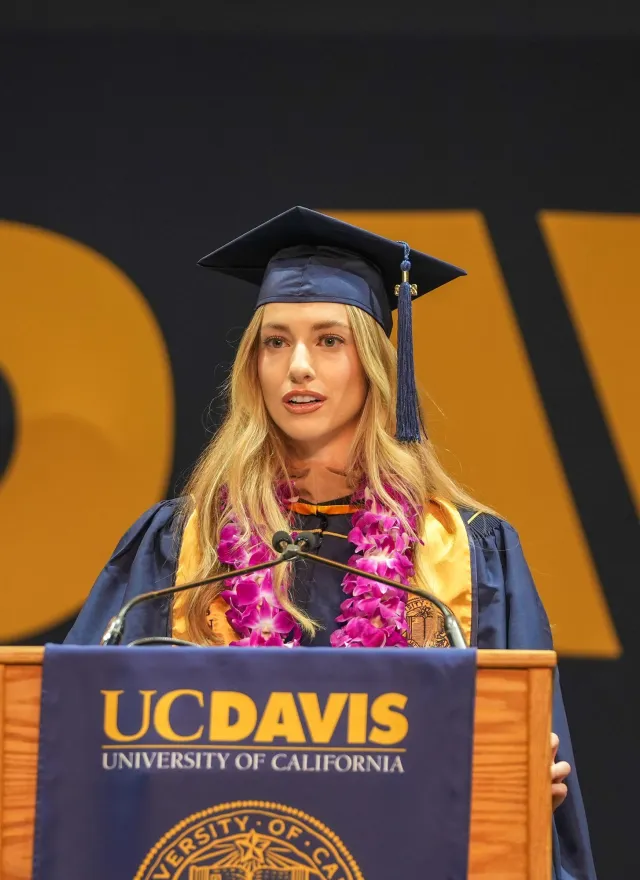 A graduate in cap and gown speaks at a podium with a UC Davis sign, wearing a purple lei, during a commencement ceremony.