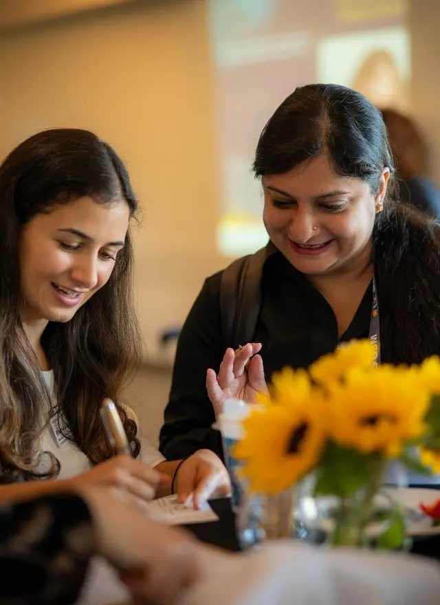 Two women sit at a table with sunflowers, smiling and writing on paper during an indoor event.