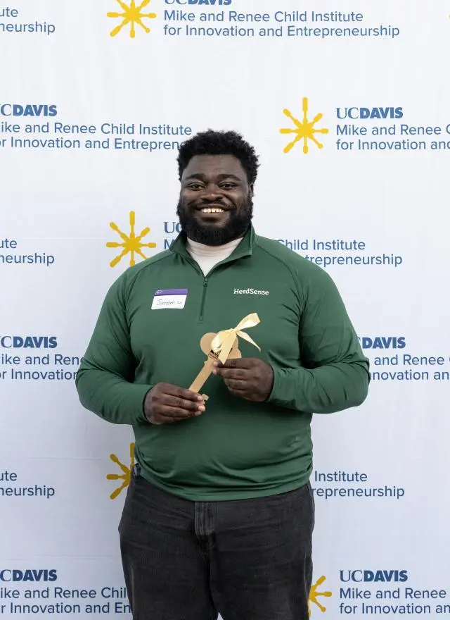 Jeremy Twumasi Ampofo in a green polo shirt holds a wooden award in front of a UC Davis backdrop.