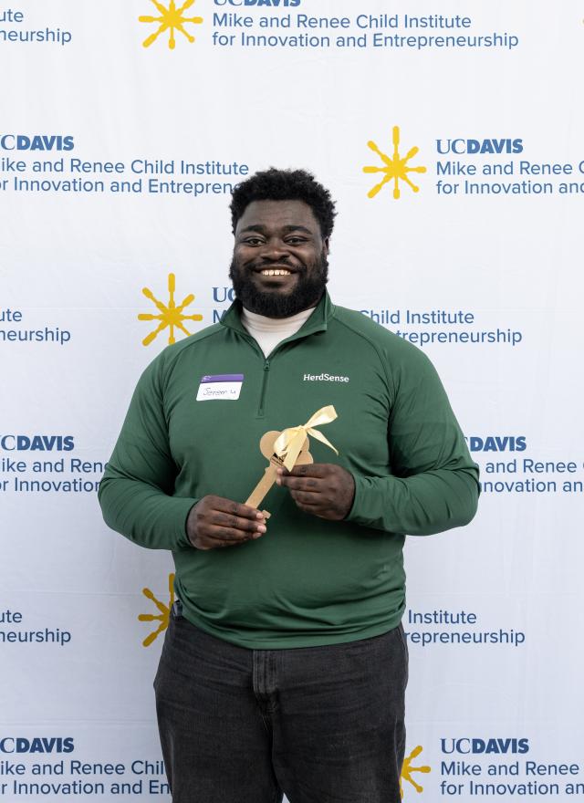 Jeremy Twumasi Ampofo in a green polo shirt holds a wooden award in front of a UC Davis backdrop.