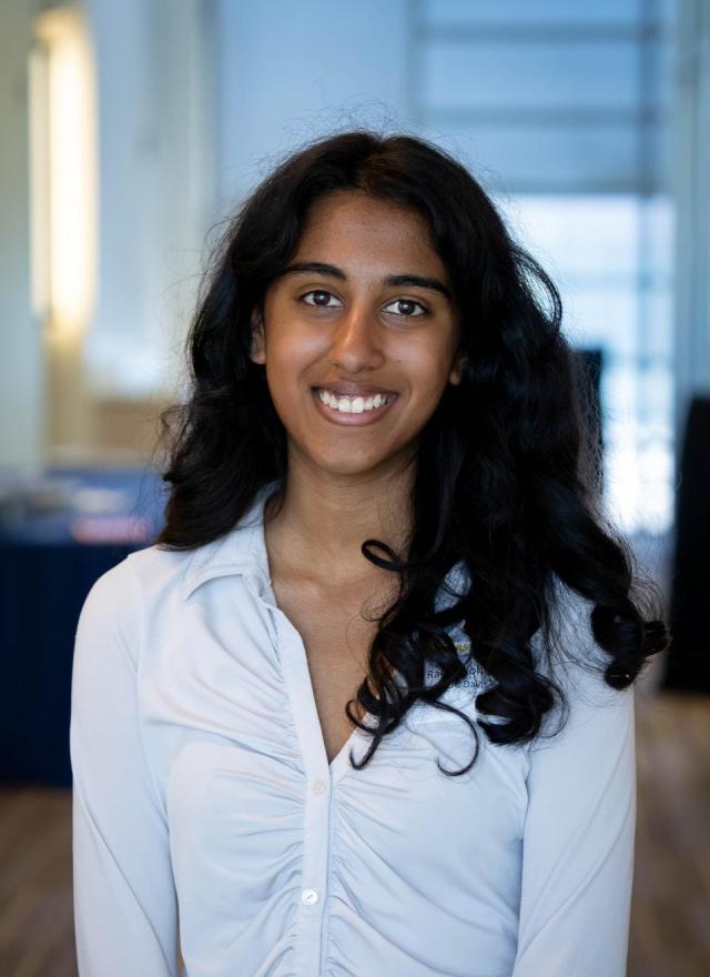 Rachel Johnson with long curly hair, wearing a white ruched blouse, stands indoors and smiles at the camera. The background is out of focus.