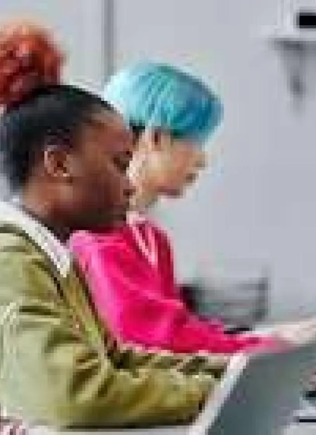 Three people are sitting in a row at desks, working on laptops in a modern classroom or office setting. Computer code is visible on a monitor in the background.