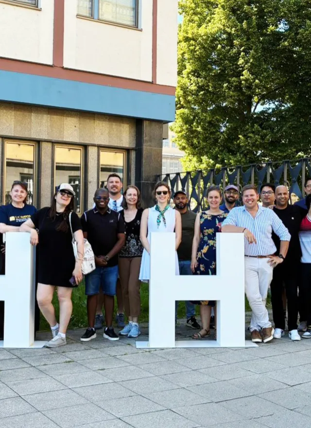 A group of people pose outdoors, smiling and standing behind large white and yellow letters spelling "HHL" on a paved area near a building and trees.