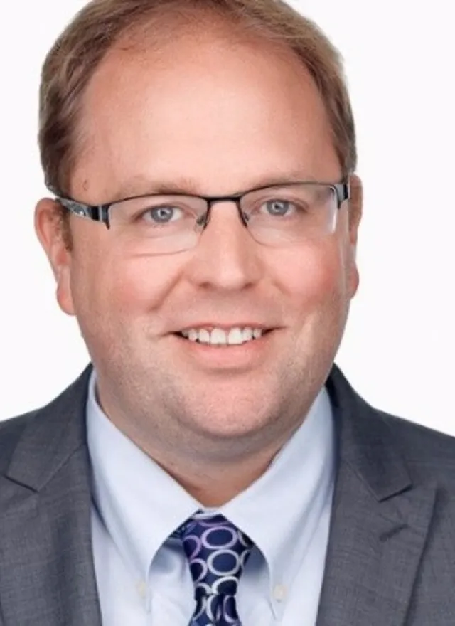 Man with short light brown hair, glasses, and a gray suit jacket over a light blue shirt and patterned tie, smiling at the camera against a plain white background.