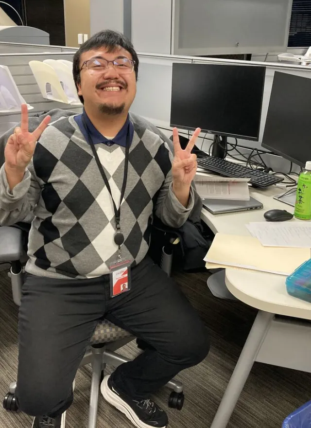 Man sitting at an office desk, smiling and making peace signs with both hands. Multiple monitors, papers, and office supplies are visible around him.