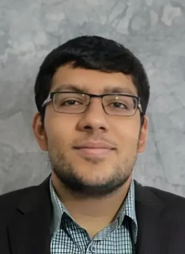A man wearing glasses and a checked shirt with a dark blazer is posed in front of a gray textured background.