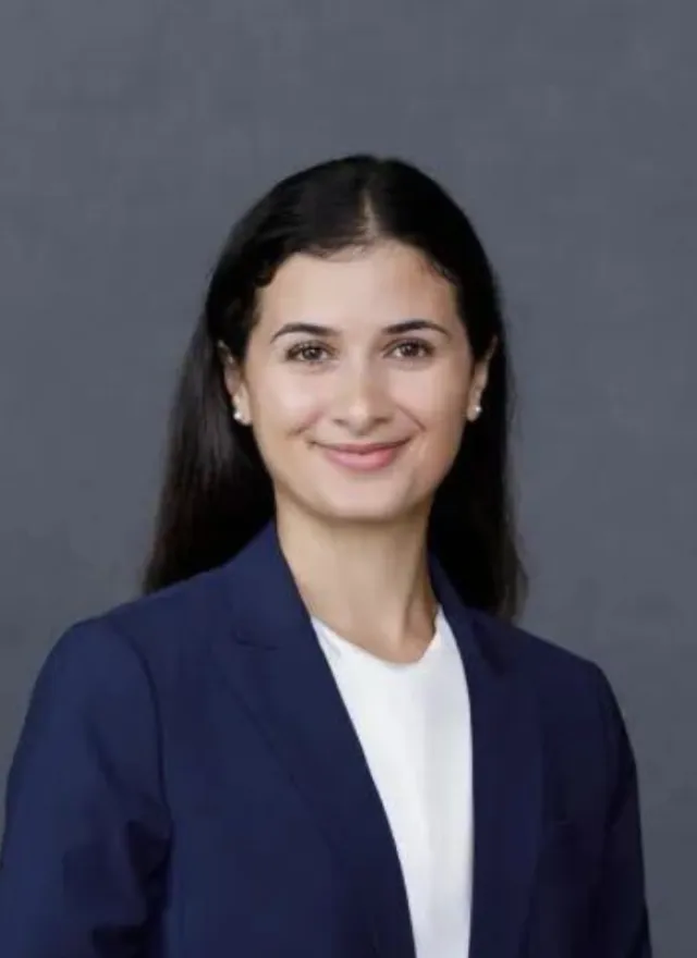 Woman with long dark hair wearing a navy blazer and white blouse, smiling in front of a plain gray background.