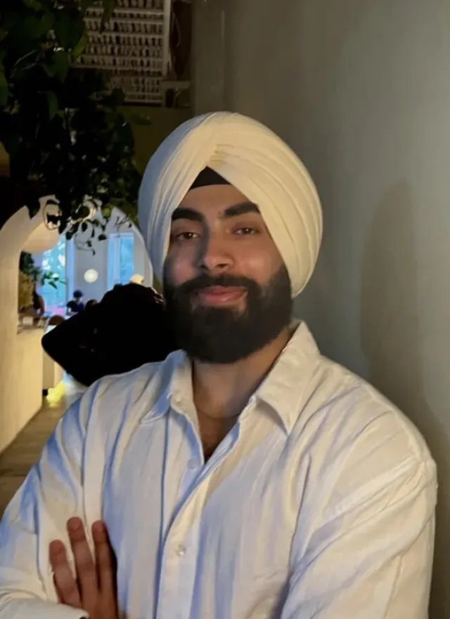 A person wearing a white turban and white shirt stands indoors with arms crossed, facing the camera and smiling slightly.