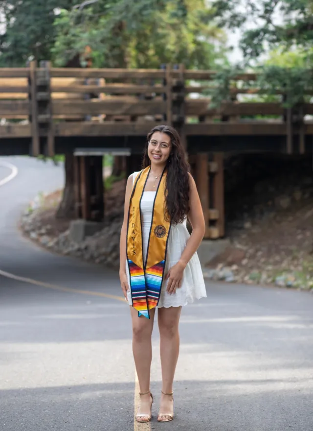 Woman in a white dress and graduation stole stands on a road in front of a wooden bridge, smiling at the camera.