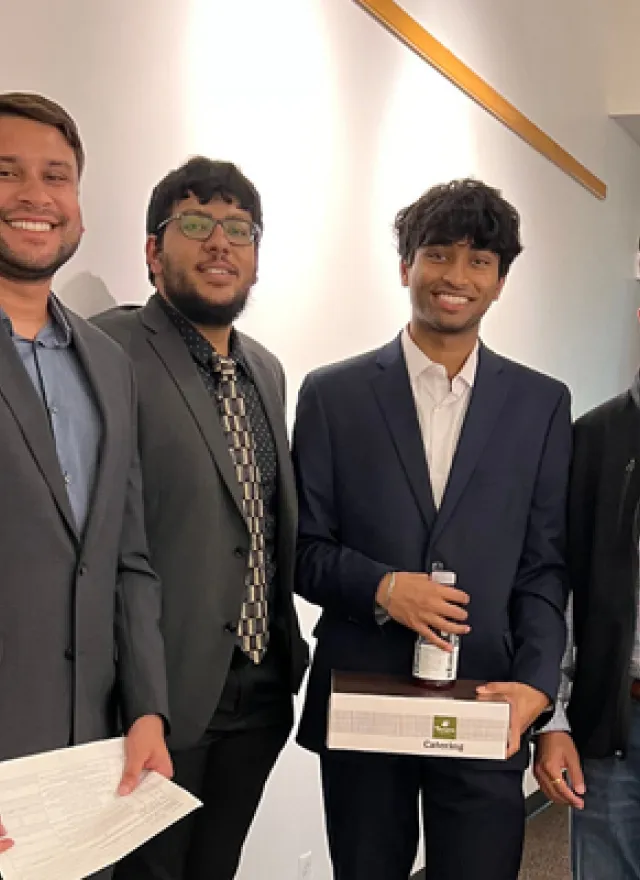 Four men in business attire stand together indoors; one holds a certificate, another holds a box and bottle. A UC Davis vest logo is visible on one person.
