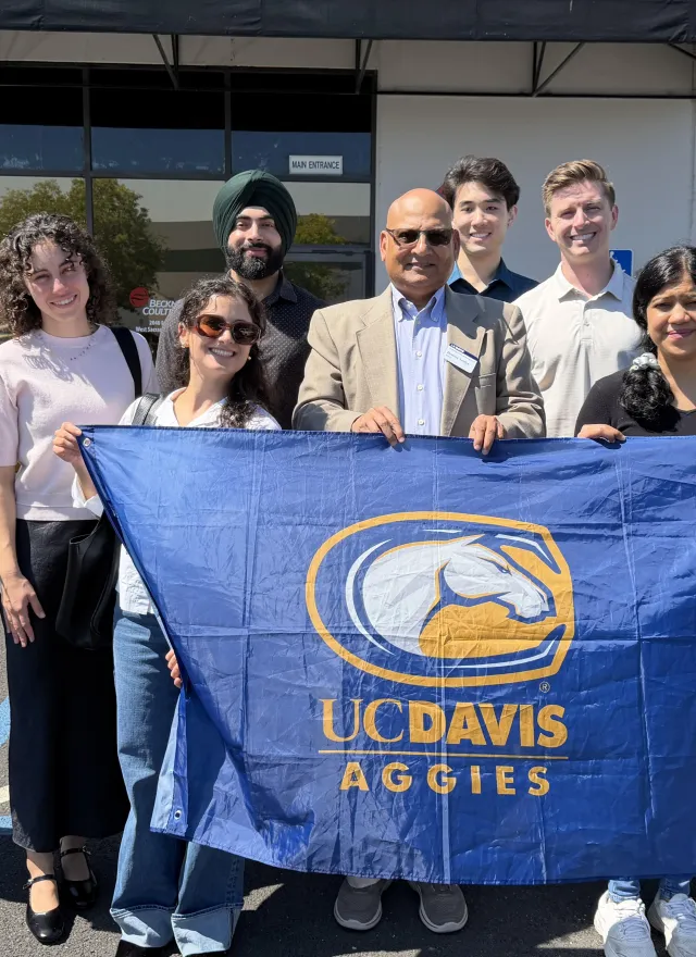 A group of people stand outdoors holding a blue UC Davis Aggies flag; one person holds a box, and an American flag is visible in the background.