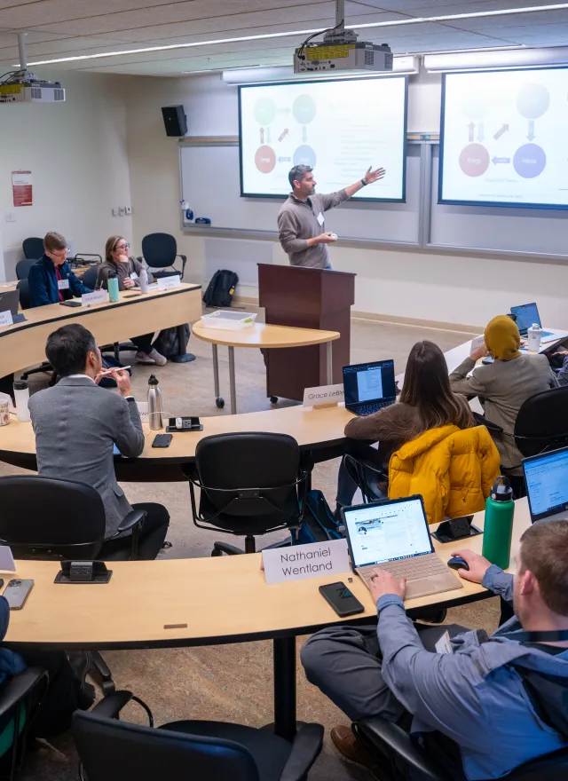 A lecturer presents to students seated in a semicircular classroom, some taking notes on laptops, with slides projected on screens at the front.