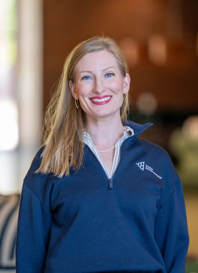 Jaimie Asakawa smiling, wearing a navy pullover and pearl necklace, standing indoors with blurred background.