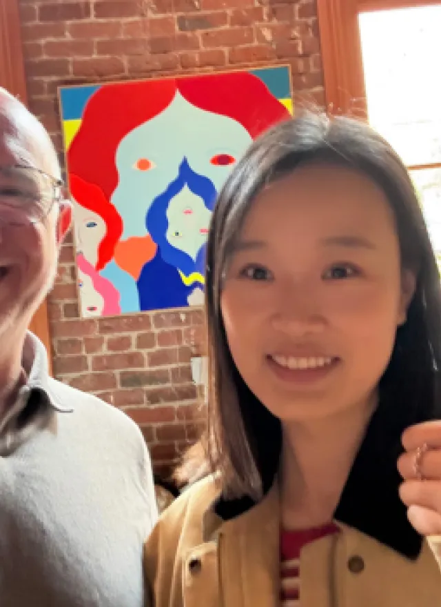 Two people smile at the camera indoors; one gives a thumbs up. A colorful abstract painting and large windows with a garden view are in the background.