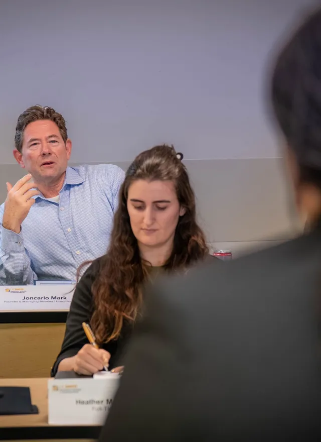 A man speaks during a classroom discussion while two women listen and take notes. Nameplates and notebooks are on the desks.
