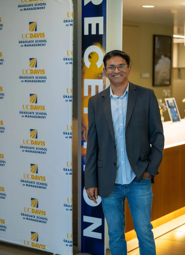 A man in a blazer and jeans stands smiling beside a UC Davis Graduate School of Management banner in an indoor setting.