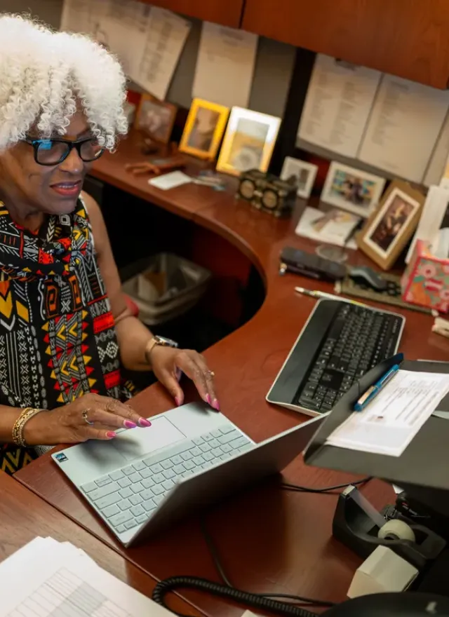 Woman with white curly hair works at a desk with a laptop and monitor, surrounded by paperwork and framed photos.