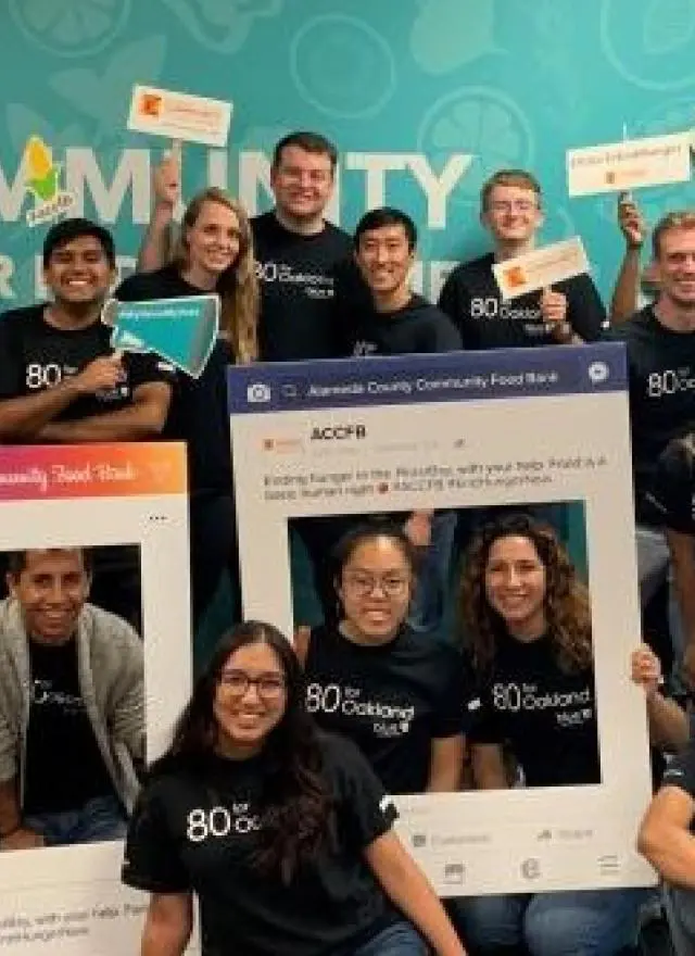 A group of people in matching black shirts pose together indoors, holding signs and frames promoting community service and volunteer work.