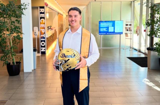 A man in business attire and a graduation stole stands holding a UC Davis football helmet in the lobby of the UC Davis Graduate School of Management.