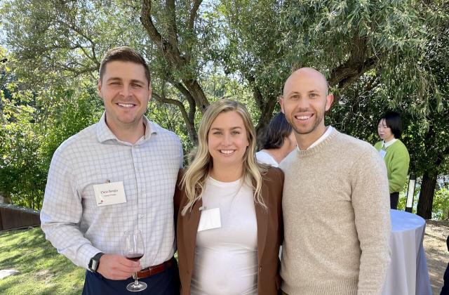 Three people stand outdoors, smiling at the camera. The two men wear business casual attire; the woman wears a white top and brown blazer. Trees and other event attendees are in the background.