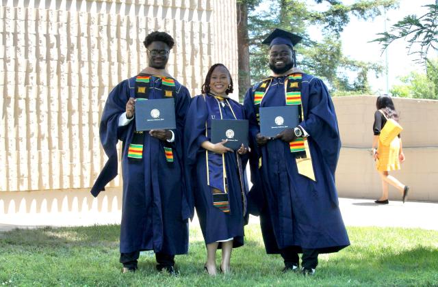 Three graduates in navy blue caps and gowns hold diplomas and smile outdoors; another person walks in the background.
