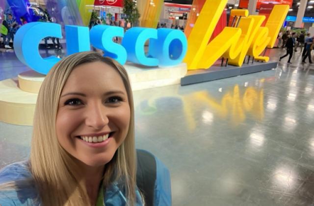 A woman takes a selfie in front of a large "CISCO Live" sign at a convention or expo, with colorful decorations and other attendees in the background.