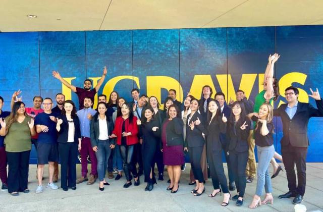 A group of people posing and smiling in front of a large blue and yellow “UC DAVIS” sign, with several making enthusiastic gestures.