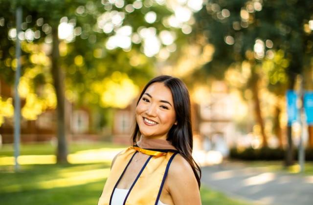 A young woman in a graduation stole and white dress stands outdoors by a brick wall, smiling, with trees and sunlight in the background.