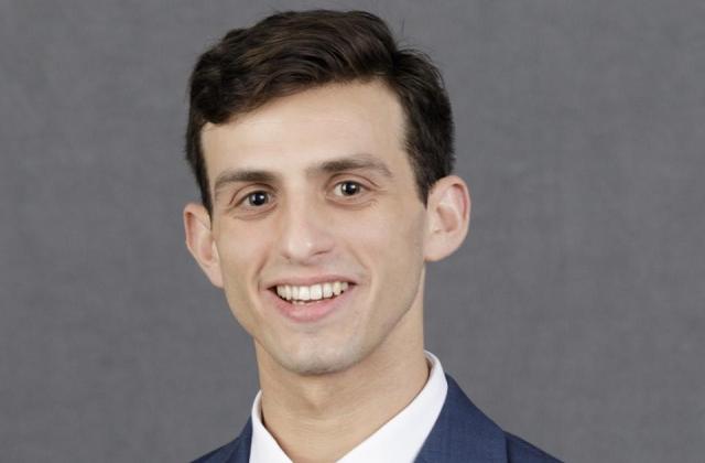 Jacob Renneisen wearing a blue suit, white shirt, and red tie is smiling in front of a plain gray background.