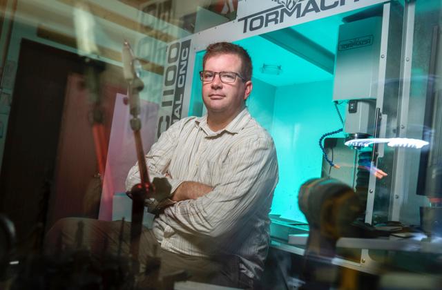 A man wearing glasses and a striped shirt sits with arms crossed in front of a Tormach CNC machine in a workshop.