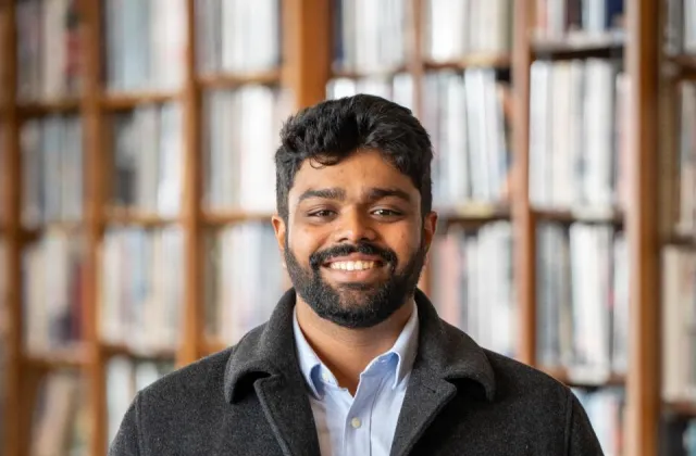 A man with dark hair and a beard, wearing a dark coat and light shirt, stands smiling in front of bookshelves filled with books.