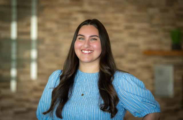Lizzie Loredo stands smiling in front of a blurred brick wall background.
