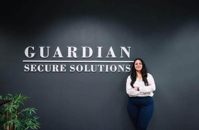 A woman stands with arms crossed in front of a dark wall displaying the words "Guardian Secure Solutions.