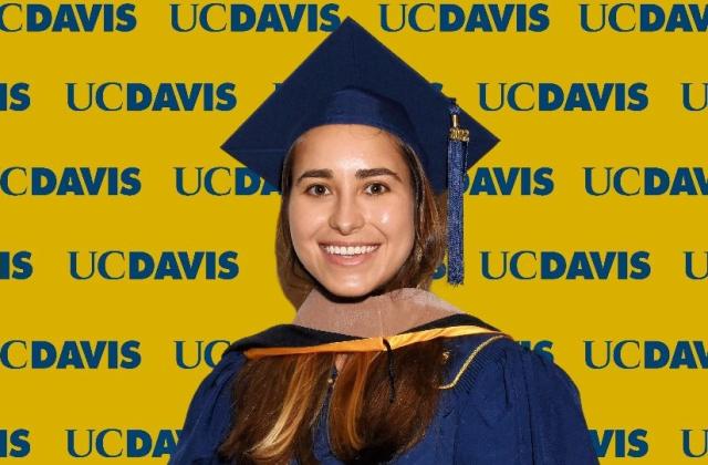 Maria Ahumada wearing a blue graduation cap and gown stands in front of a yellow UC Davis backdrop with repeated logos.