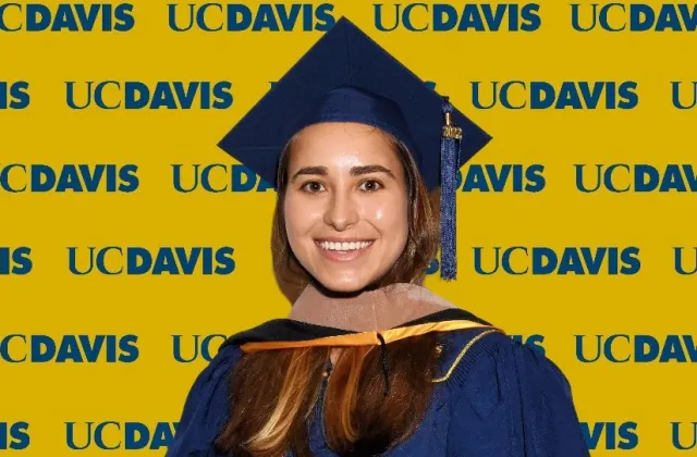 Maria Ahumada wearing a blue graduation cap and gown stands in front of a yellow UC Davis backdrop with repeated logos.