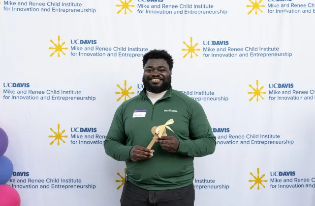 Jeremy Twumasi Ampofo in a green polo shirt holds a wooden award in front of a UC Davis backdrop.