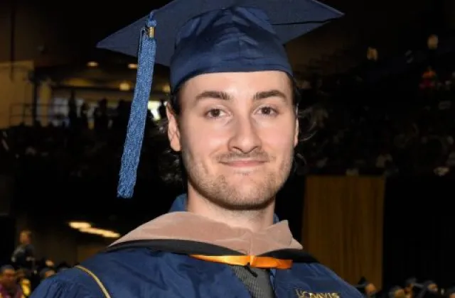 A person in a blue graduation cap and gown stands indoors at a graduation ceremony, with other graduates and an audience visible in the background.