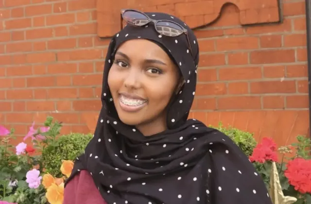 A woman wearing a black and white polka dot hijab and sunglasses on her head smiles in front of a brick wall and colorful flowers.