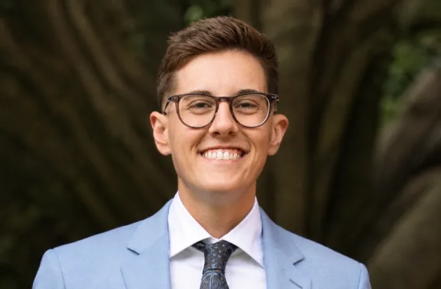 A person wearing glasses and a light blue suit jacket smiles at the camera, standing outdoors with a tree trunk in the blurred background.