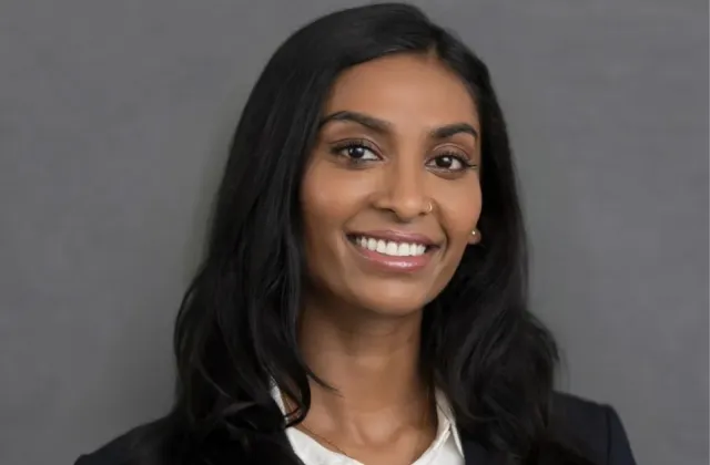 Woman with long dark hair, wearing a white shirt and dark blazer, smiling in front of a plain gray background.
