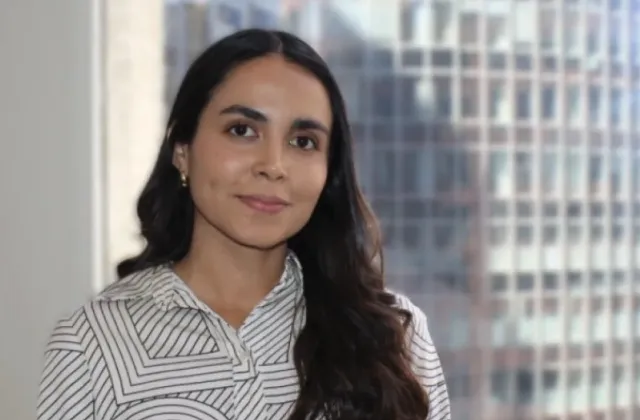 A woman with long dark hair wearing a patterned blouse stands in front of a window with a cityscape in the background.