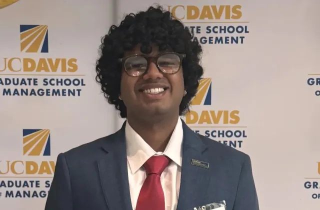 Person in a suit, red tie, and glasses smiling in front of a UC Davis Graduate School of Management backdrop.
