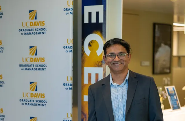 A man in a blazer and jeans stands smiling beside a UC Davis Graduate School of Management banner in an indoor setting.
